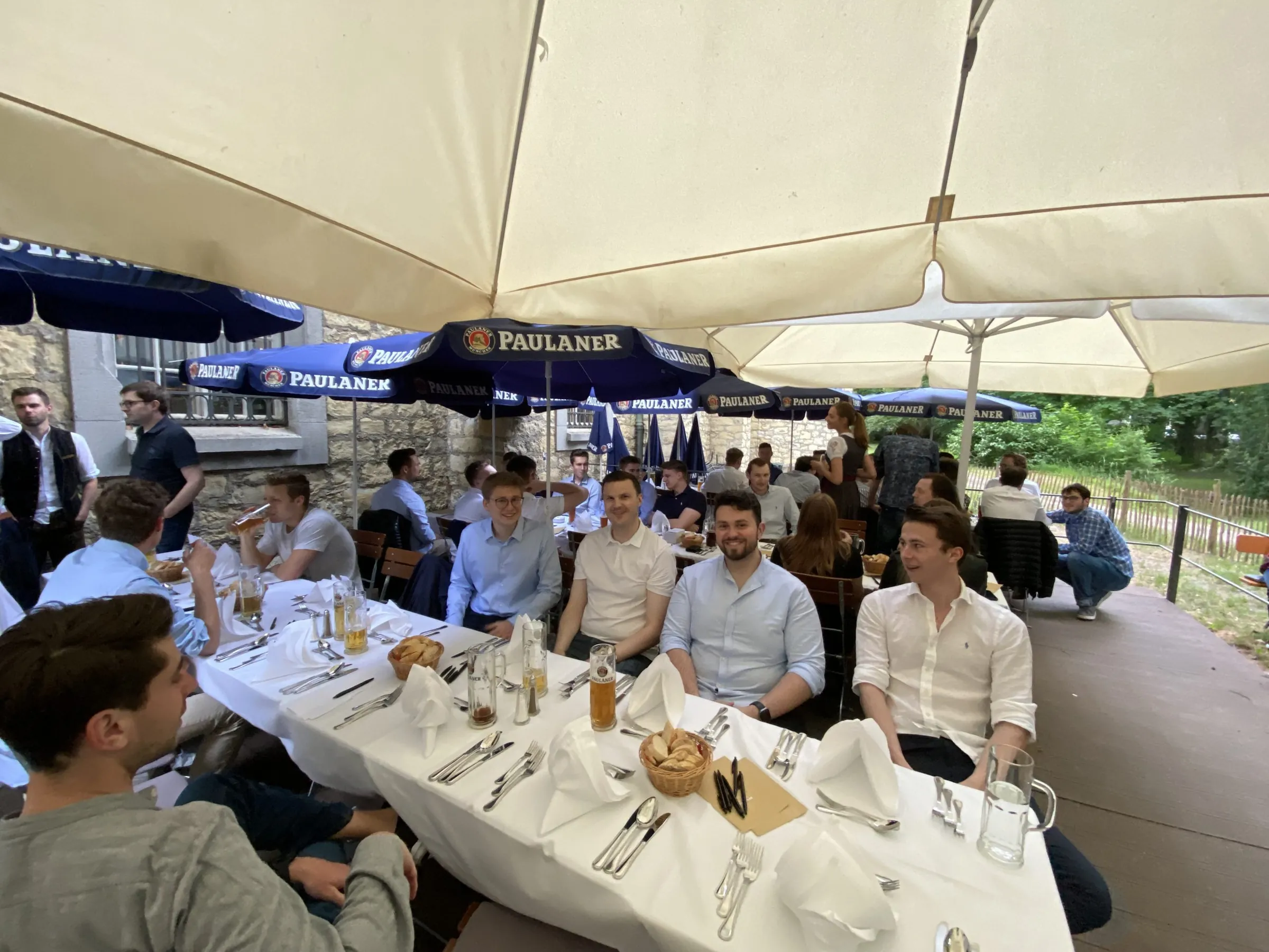 Mitglieder des Investment Club Regensburg an einer langen, weiß gedeckten Tafel in einem Biergarten unter Sonnenschirmen.