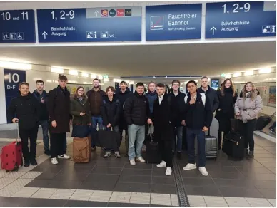 Gruppe von Studierenden mit Koffern und Taschen in einer Bahnhofshalle unter den Gleisanzeigern.