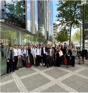 Große Gruppe von Studierenden in Business-Kleidung steht auf einem Gehweg vor einem verglasten Bürogebäude.