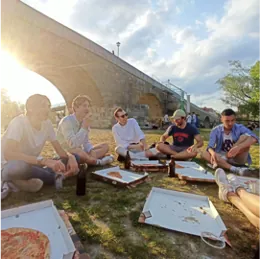 Studierende sitzen im Gras auf einer Wiese und essen Pizza, im Hintergrund eine steinerne Bogenbrücke im Gegenlicht.