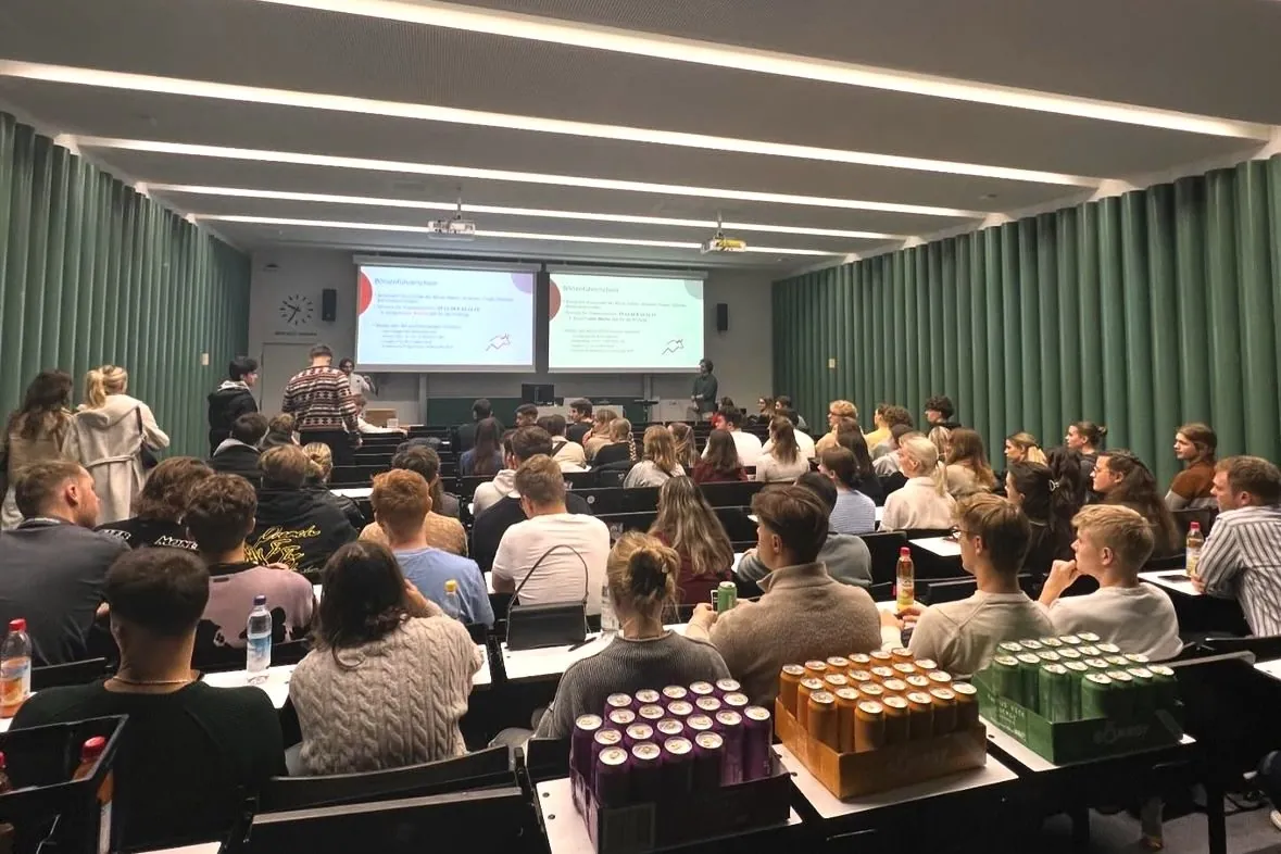 Gut gefüllter Hörsaal bei einer Veranstaltung des Investment Club Regensburg, vorne die Präsentation auf zwei Leinwänden, auf den Tischen Getränkedosen.